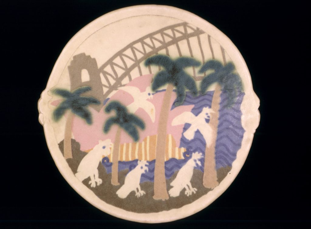 Rounded stoneware platter, unfooted flat base flaring to a shallow bowl with two small protruding handles at each side. The platter features a light coloured stoneware body, decorated under the glaze with a Sydney harbour scene, the Opera House (in pink), palm trees in grey and blue and reserve colouredcockatoos in the foreground with the Sydney Harbour Bridge painted in grey in the background. An undecorated border frames the image. The glaze is a light grey/buff, crackled and opaque in parts for decorative effect.